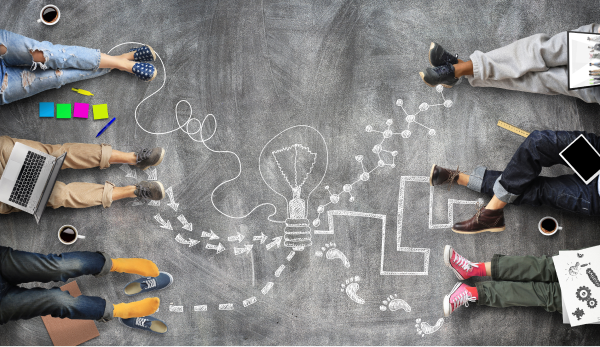 Overhead view of a collaborative team brainstorming on a chalkboard floor with creative doodles, colorful sticky notes, and technology.