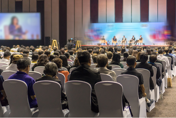 A large audience attending a professional conference in a spacious hall, watching a panel discussion on stage. The stage is brightly lit, with multiple speakers seated and engaged in conversation, while the audience listens attentively.